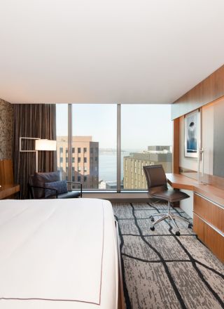 A modern hotel room with a large bed, wide TV, desk, and city view from a window, featuring stylish decor and a comfortable atmosphere.