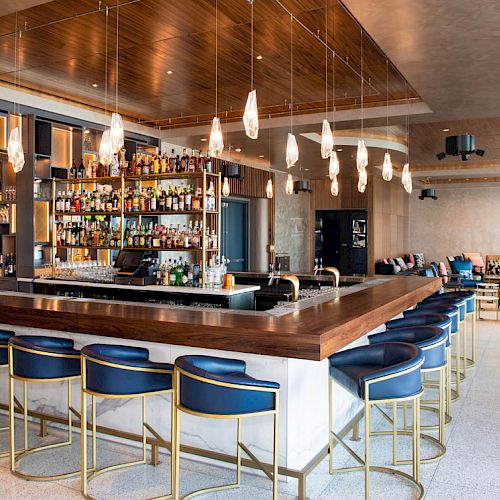 A modern bar with blue stools, pendant lights, and a stocked liquor shelf; cozy seating in a spacious, sophisticated setting.
