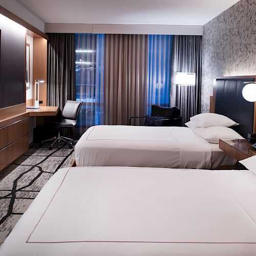 The image shows a modern hotel room with two neatly made beds, a wall-mounted TV, a desk, and stylish lighting, featuring contemporary decor.