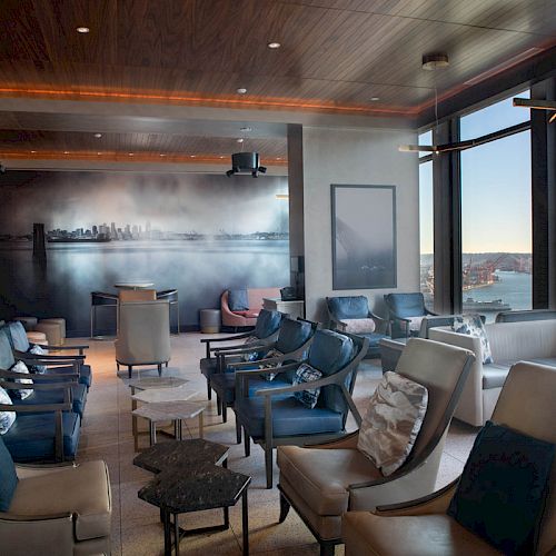 The image shows a modern lounge area with comfortable seating, large windows offering a scenic view of a cityscape and waterfront.