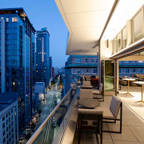 A modern urban rooftop terrace at dusk, overlooking city skyscrapers, with tables and seating along the balcony railing.