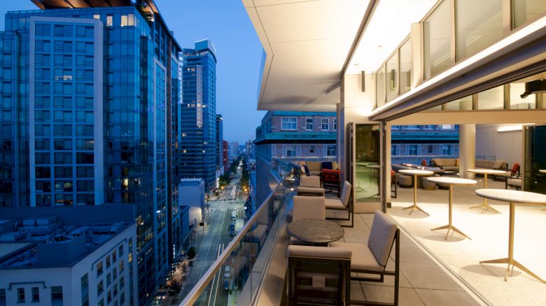 Rooftop terrace with seating and tables, overlooking a cityscape with tall buildings along a lit street at dusk.