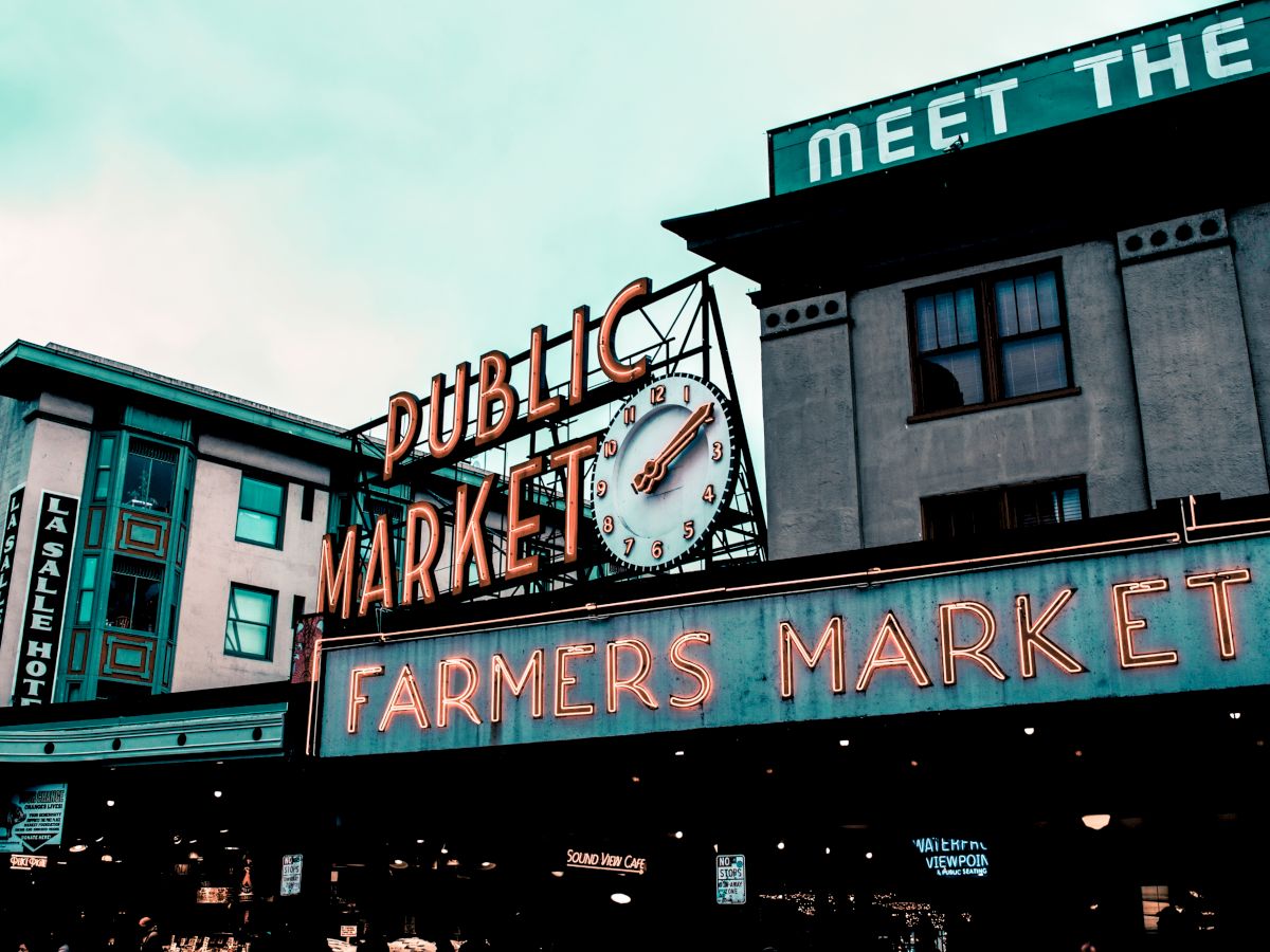 The image shows the iconic neon signs of a public and farmers market, with a clock, set against a backdrop of buildings in a bustling area.