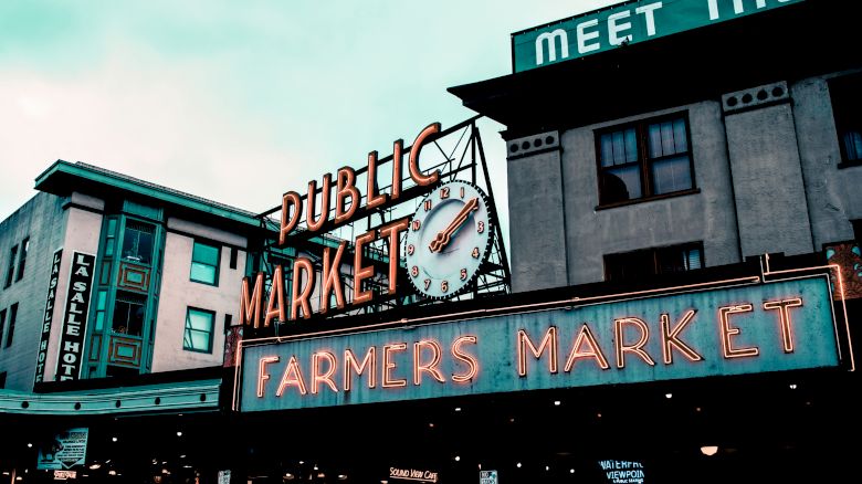The image shows the iconic neon signs of a public and farmers market, with a clock, set against a backdrop of buildings in a bustling area.