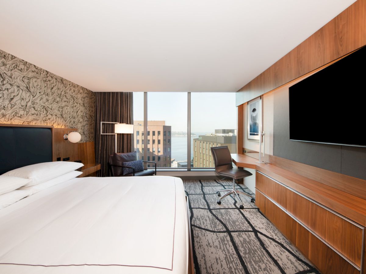 A modern hotel room features a large bed, a wall-mounted TV, a desk, and a view of buildings and water through a window.