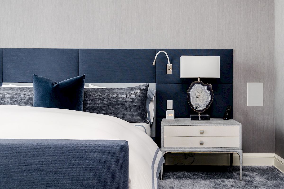 A modern bedroom featuring a bed with navy headboard, grey pillows, and a white nightstand with a lamp, set on plush carpet flooring.