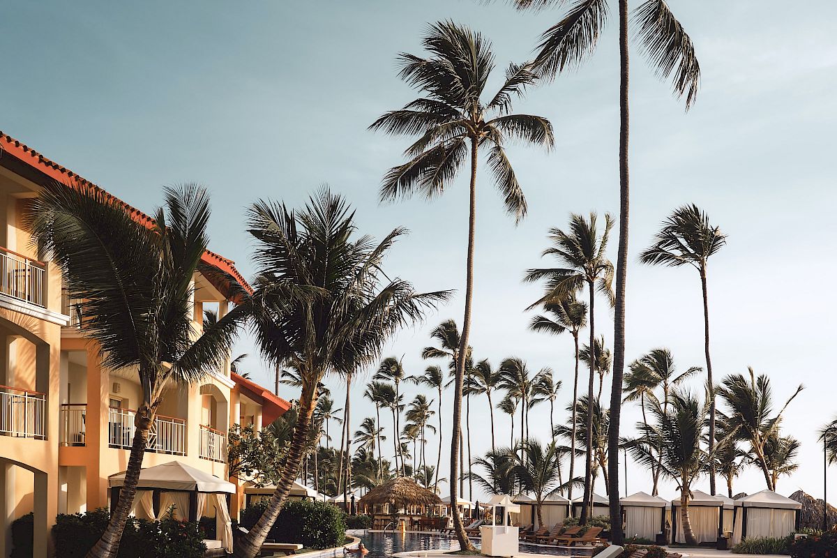 A tropical resort scene with palm trees, a swimming pool, lounge chairs, and a building. The sky is clear, creating a relaxing atmosphere.