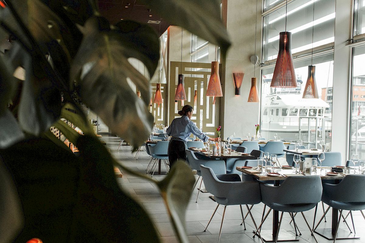 A person is setting tables in a stylish, modern restaurant with hanging lights and large windows, partially obscured by plants.