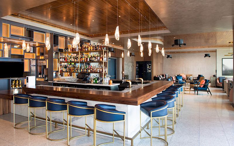 A modern bar with blue stools, suspended lights, and a fully stocked back bar. The space is open with additional seating and decor elements.