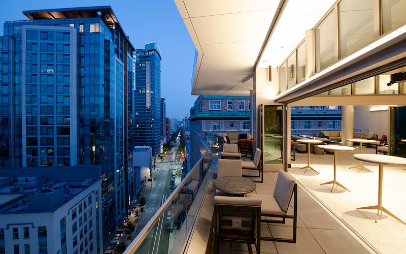 A modern rooftop restaurant with outdoor seating overlooks a city street lined with tall buildings, captured at twilight.