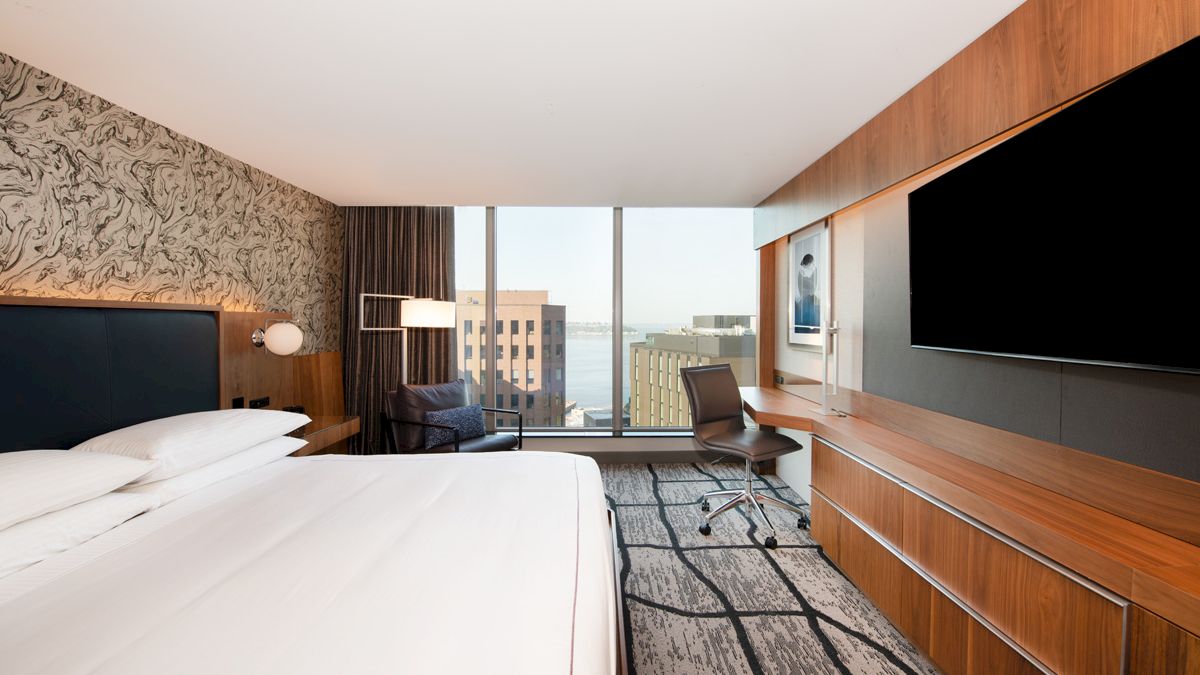 A modern hotel room with a large bed, desk, chair, TV, and a view of city buildings through the window is shown.