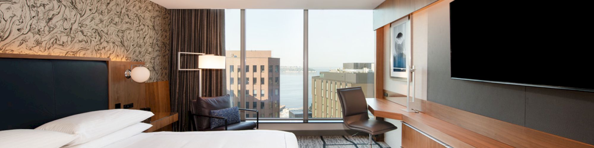 A modern hotel room with a large bed, desk, chair, TV, and a view of city buildings through the window is shown.