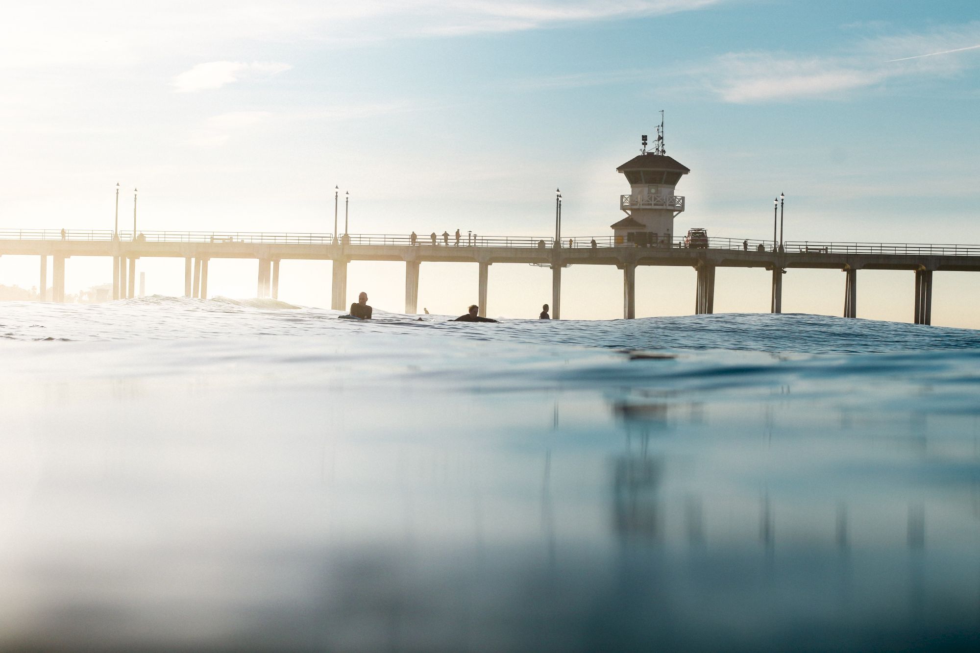 A serene ocean view with surfers in the foreground and a long pier extending into the water, under a clear sky.